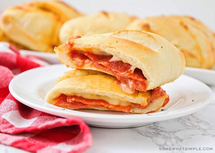 a homemade pepperoni pizza pocket that has been cut in half and the two halves are stacked on top of each other. In the background is a tray filled with several more golden brown pizza pockets.