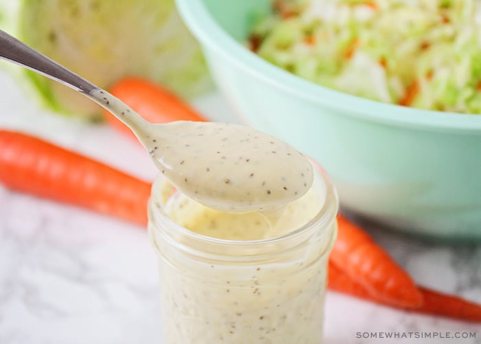 a jar of homemade coleslaw dressing with a spoon above the jar filled with dressing. A bowl of coleslaw in a blue bowl is in the background