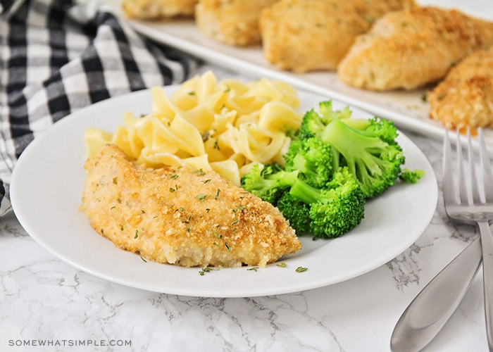 a white plate with a breast of baked garlic parmesan chicken with a side of pasta and broccoli. In the background there are five more breasts of garlic parmesan chicken