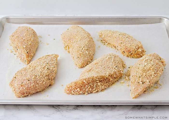 a baking tray lined with parchment paper with six raw chicken breasts that have been covered in bread crumbs