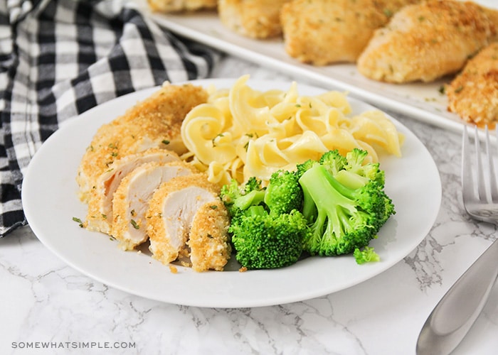 a plate with a breast of garlic parmesan chicken that has been sliced with a side of pasta noodles and broccoli next to it on the plate. A tray of additional parmesan chicken breasts are in the background.