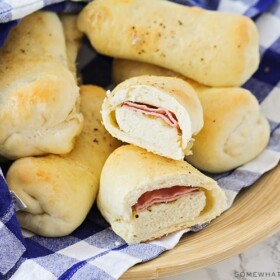 a bowl of baked Brazilian ham and cheese rolls that are golden brown and wrapped in a blue and white checkered cloth napkin. One of the rolls has been cut in half and you can see the smoked ham and mozzarella cheese inside.