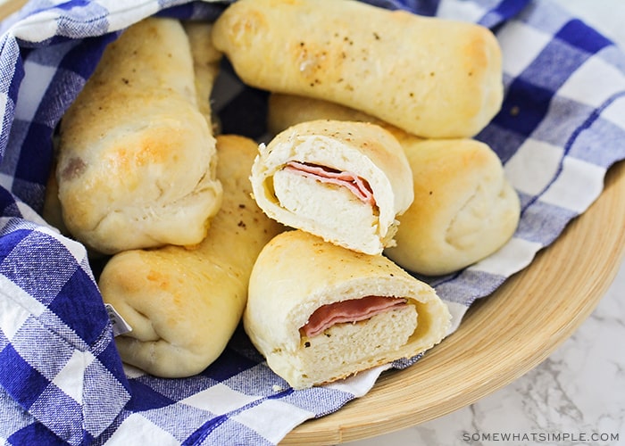 a bowl of baked Brazilian ham and cheese rolls that are golden brown and wrapped in a blue and white checkered cloth napkin. One of the rolls has been cut in half and you can see the smoked ham and mozzarella cheese inside.