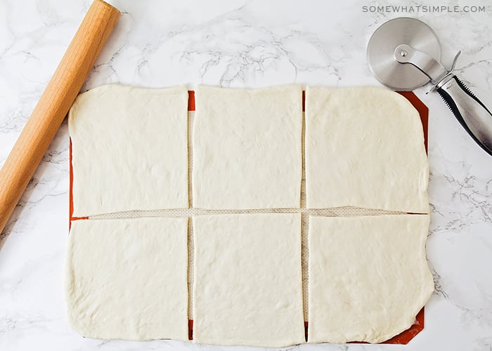 raw dough spread out on a counter and cut into six equal pieces. A wooden rolling pin and a pizza cutter are laying next to the dough on the counter.