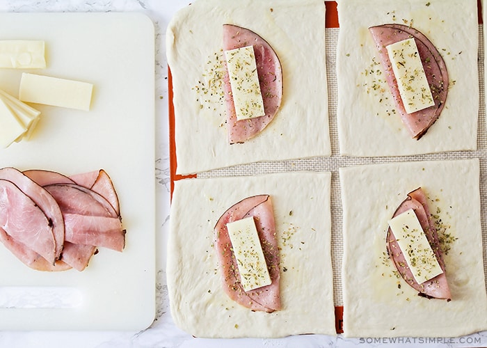 squares of dough that have a slice of smoked ham and a slice of mozzarella cheese, topped with oregano. Next to the dough is a stack of ham and cheese on a white cutting board.