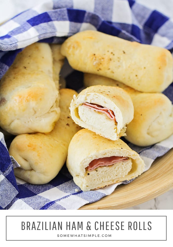 a close up of bowl of baked Brazilian ham and cheese rolls that are golden brown and wrapped in a blue and white checkered cloth napkin. One of the rolls has been cut in half and you can see the smoked ham and mozzarella cheese inside. At the bottom of the image, the words Brazilian ham & cheese rolls is written in black letters.
