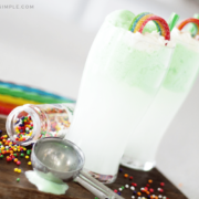 two green st. patrick's day drinks on a cutting board topped with a rainbow candy and a green piece of licorice for a straw. There are colored sprinkles spilled on the cutting board with an ice cream scoop laying next to the drink and colored pieces of licorice in the background.