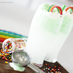 two green st. patrick's day drinks on a cutting board topped with a rainbow candy and a green piece of licorice for a straw. There are colored sprinkles spilled on the cutting board with an ice cream scoop laying next to the drink and colored pieces of licorice in the background.