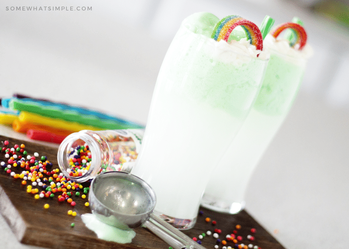 two shamrock floats on a cutting board topped with a rainbow candy and a green piece of licorice for a straw. There are colored sprinkles spilled on the cutting board with an ice cream scoop laying next to the drink and colored pieces of licorice in the background.