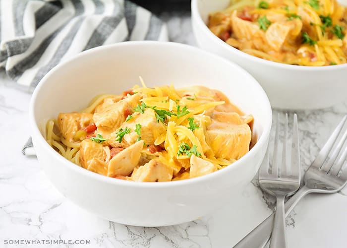 two white bowls filled with crock pot cheesy chicken spaghetti sitting on a counter. Next to the bowls are two forks in an X pattern and a black and white striped towel.