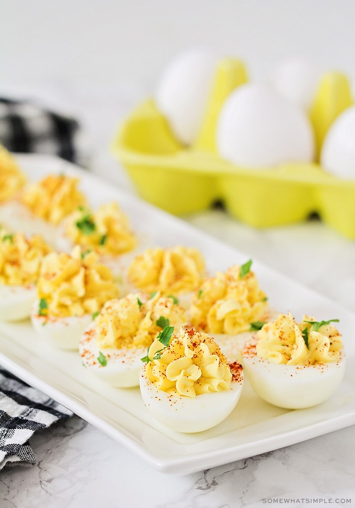 a serving tray of classic deviled eggs that are filled with a paste made of Dijon mustard and mayonnaise and topped with paprika and shredded parsley. A yellow carton of eggs is in the background.