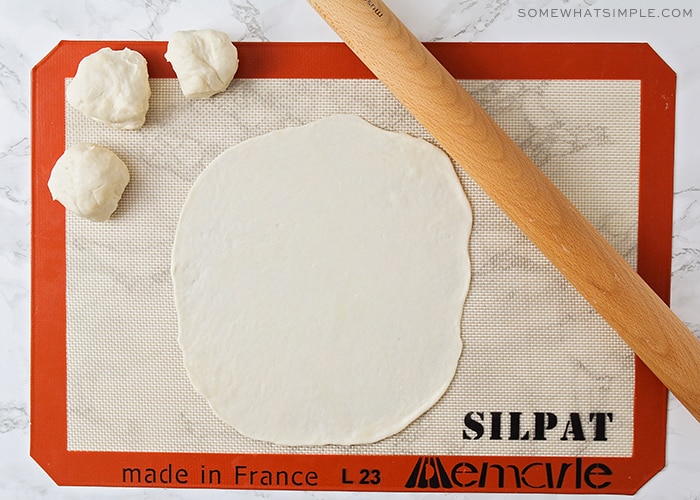 a piece of dough that has been rolled out on a baking mat. Next to the flat piece of dough are three balls of dough and on the other side is a wooden rolling pin