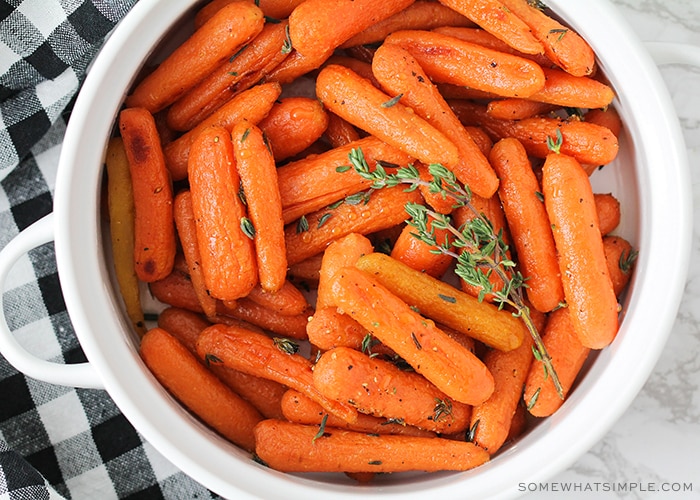 looking down on a white bowl filled with oven roasted honey glazed carrots topped with thyme. A black and white checkered napkin is laying next to the bowl on the counter.