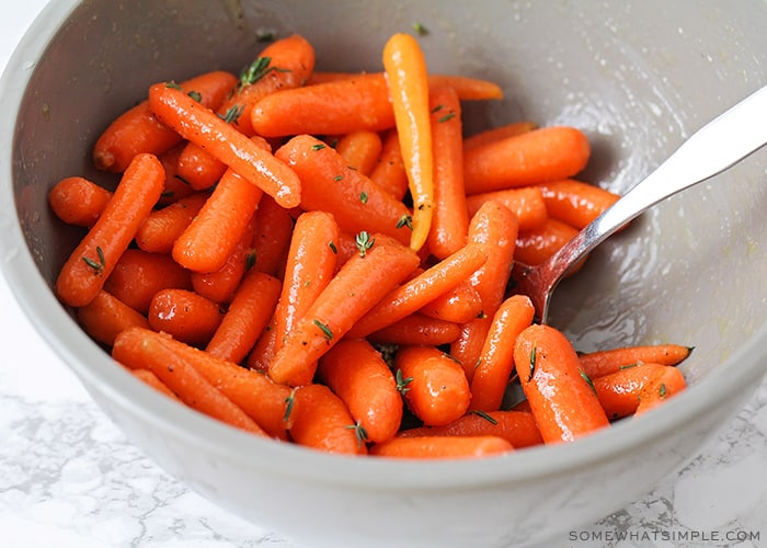 a gray bowl filled with baby carrots. A spoon is in the bowl and the carrots have just been tossed in a honey and olive oil mixture and thyme has been sprinkled over the top.