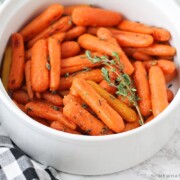 a white serving bowl that is filled with honey glazed carrots that have been baked and topped with thyme. A black and white checkered napkin is laying next to the bowl on the counter.