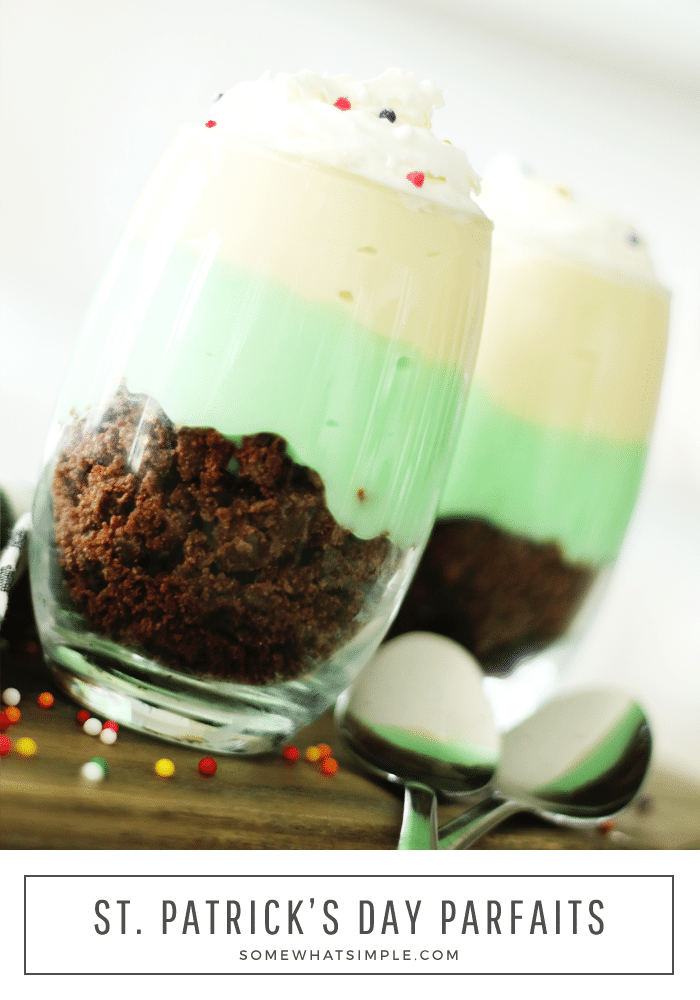 a close up of two glass cups of pistachio parfaits that are filled with brownie crumbs at the bottom, followed by a layer of pistachio pudding and a layer of vanilla pudding on top. Both cups are topped with whipped cream and round sprinkles. The cups are sitting on a wood cutting board and two spoons are laying next to the parfait cups and round sprinkles are scattered on the cutting board. The words st. Patrick's day parfaits are written at the bottom of the image in a white box.