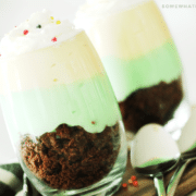 two glass cups of pistachio parfaits that are filled with brownie crumbs at the bottom, followed by a layer of pistachio pudding and a layer of vanilla pudding on top. Both cups are topped with whipped cream and round sprinkles. The cups are sitting on a wood cutting board and two spoons are laying next to the parfait cups.