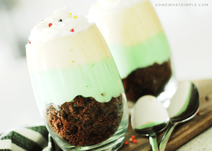 two glass cups of pistachio parfaits that are filled with brownie crumbs at the bottom, followed by a layer of pistachio pudding and a layer of vanilla pudding on top. Both cups are topped with whipped cream and round sprinkles. The cups are sitting on a wood cutting board and two spoons are laying next to the parfait cups.