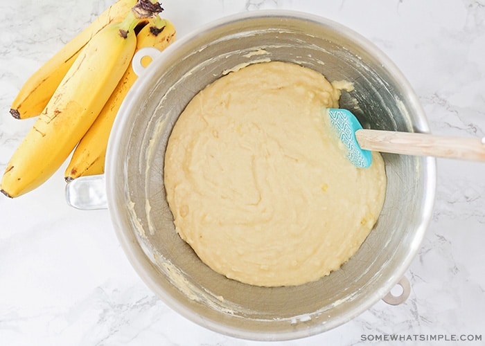 Looking down on a metal bowl filled with banana bread batter and a blue spatula inside. Next to the bowl is a bunch of three bananas.