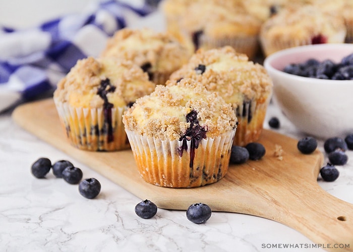 four baked blueberry muffins with a cinnamon crumb topping are sitting on a wood cutting board. Next to the muffins is a bowl of fresh blueberries.