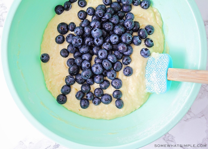 looking down on a light blue mixing bowl filled with fresh blueberries and batter with a spatula in the bowl laying against the side.