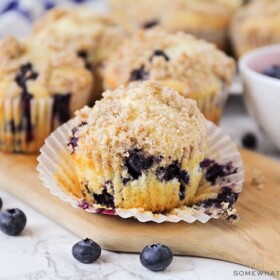 a close up of a blueberry muffin that has the cupcake liner pulled down. Behind the muffin are additional muffins as well as a bowl of fresh blueberries.