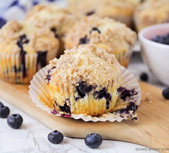 a close up of a blueberry muffin that has the cupcake liner pulled down. Behind the muffin are additional muffins as well as a bowl of fresh blueberries.