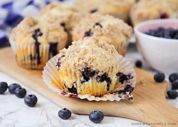 a close up of a blueberry muffin that has the cupcake liner pulled down. Behind the muffin are additional muffins as well as a bowl of fresh blueberries.