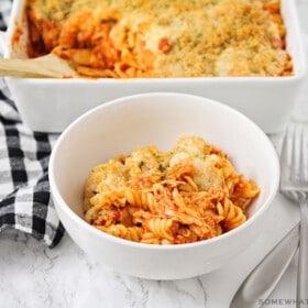 a white bowl filled with a serving of chicken parmesan casserole that is made with pasta and topped with bread crumbs and cheese. Behind the bowl on the counter is a casserole pan filled with the remaining casserole.