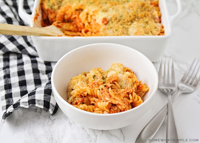 a white bowl filled with a serving of chicken parmesan casserole that is made with pasta and topped with bread crumbs and cheese. Behind the bowl on the counter is a casserole pan filled with the remaining casserole.