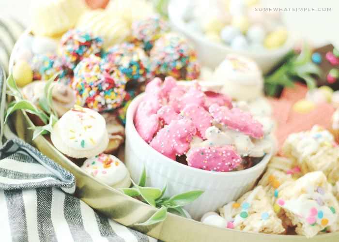an Easter cookie charcuterie board filled with several different types of cookies. There are pink and white animal cookies in a small white bowl, small sugar cookies with white frosting and pastel sprinkles, chocolate covered cookies with colorful sprinkles and other desserts in the background on the cookie tray.