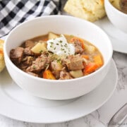 a white bowl filled with savory crock pot beef stew. There are chunks of beef and chopped potatoes and carrots topped with a dollop of sour cream and chopped parsley. The bowl is sitting on a white plate with a square of bread next to it. Above the bowl is a black and white checkered cloth napkin and in the corner of the image another bowl of stew is partially visible.