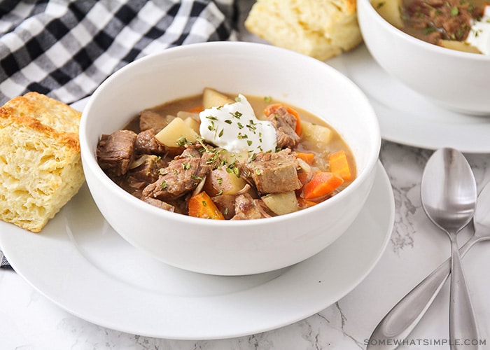 a white bowl filled with savory crock pot beef stew. There are chunks of beef and chopped potatoes and carrots topped with a dollop of sour cream and chopped parsley. The bowl is sitting on a white plate with a square of bread next to it. Above the bowl is a black and white checkered cloth napkin and in the corner of the image another bowl of stew is partially visible.