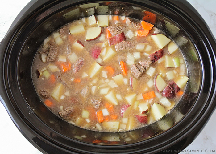 looking down on a crock pot filled with broth, beef, and chopped carrots, potatoes and other vegetables.