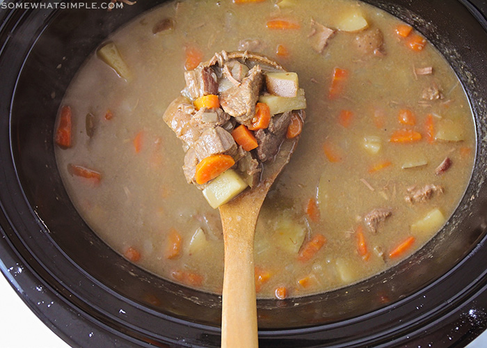 looking down on a slow cooker filled with beef stew. A wooden spoon is in the center holding chunk of beef, carrots and potatoes.