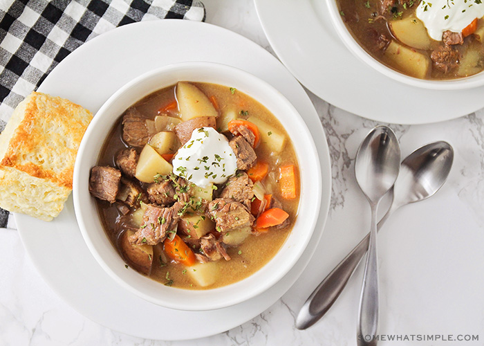 looking down on a bowl of slow cooker beef stew topped with a dollop of sour cream. A piece of bread is next to the bowl and two spoons are on the counter on the other side of the bowl.