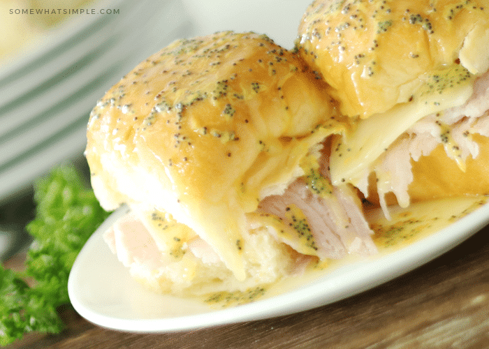 two baked ham and cheese sliders topped with butter and poppy seeds on a white plate with a pinch of parsley behind the plate.