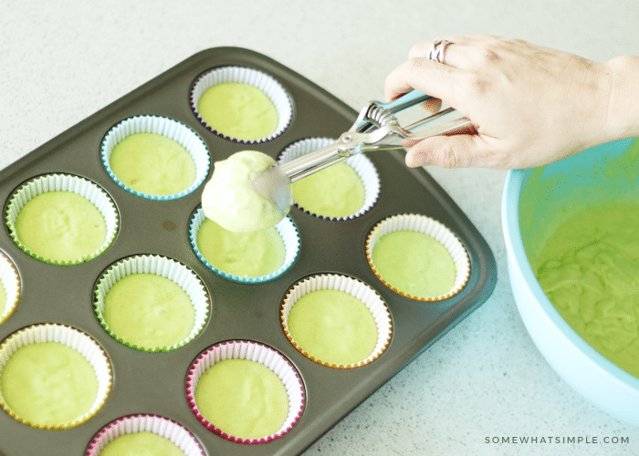 a hand holding an ice cream scoop above a cupcake tin filled with cupcake liners and raw batter in each cup. Next to the pan is a blue bowl that has key lime cupcake batter inside.