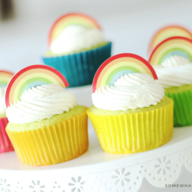 a close up of Key Lime Cupcakes sitting on top of a white cake stand. The lime cupcakes are wrapped in different colored cupcake wrappers and topped with white frosting and a rainbow cupcake topper