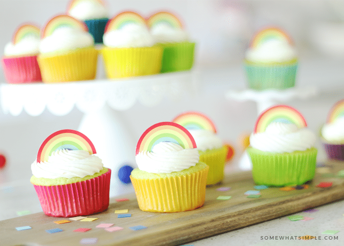 four key lime cupcakes sitting on a wood cutting board. Each are topped with white frosting and a rainbow cupcake topper. The cupcakes have different colored cupcake liners and multi colored confetti is laying on the cutting board. In the background more lime cupcakes with rainbow toppers are sitting on a white cake stand.