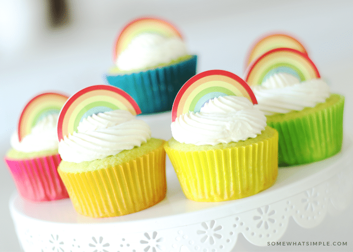 Key Lime Cupcakes sitting on top of a white cake stand. The lime cupcakes are wrapped in different colored cupcake wrappers and topped with white frosting and a rainbow cupcake topper