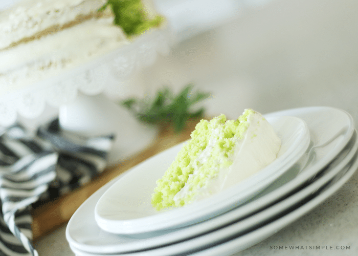a slice of lime cake on a stack of white plates with rest of the lime cake sitting on a white cake stand in the background.