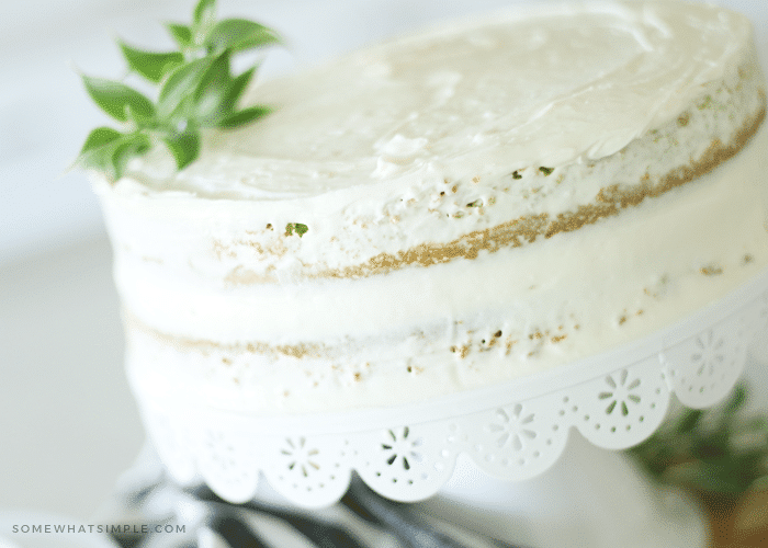 a close up of a key lime cake topped made with this easy lime cake recipe sitting on top of a white cake stand. A black and white striped cloth napkin is laying on the counter beneath the cake stand.