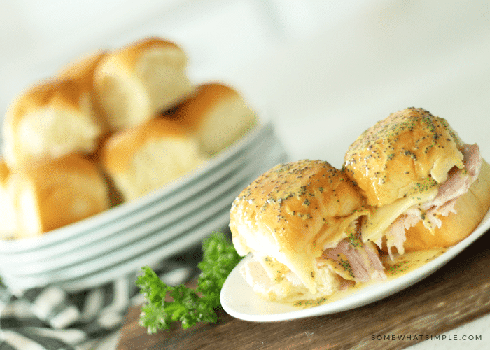 a small white plate with two ham and Swiss cheese sliders on the plate that are topped with butter and poppy seeds. Behind the plate are a stack of five white plates with a pile of King's Hawaiian rolls on top.