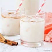 two glasses filled with this easy horchata recipe. Each glass has a red and white striped straw in it. Next to the glasses are 4 cinnamon sticks laying on the counter as well as a light red cloth napkin.