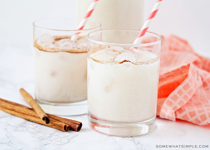 two glasses filled with this easy horchata recipe. Each glass has a red and white striped straw in it. Next to the glasses are 4 cinnamon sticks laying on the counter as well as a light red cloth napkin.