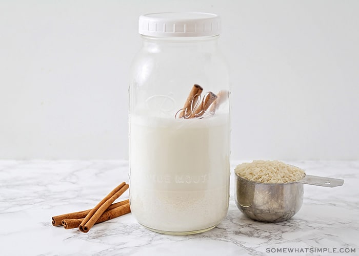 a Mason jar filled most of the way with milk with two cinnamon sticks inside. Next to the jar is a measuring cup filled with rice and a few additional cinnamon sticks.