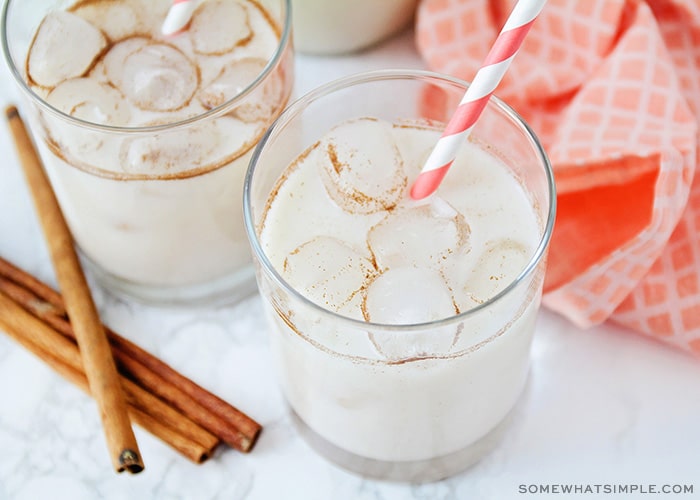 looking down on two glass filled with horchata and ice. Each cup has a striped straw inside and there are cinnamon sticks laying on the counter next to the glasses.