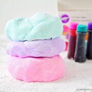 three flattened balls of marshmallow fondant stacked on top of each other on a counter covered with powdered sugar. The ball of fondant on top is light blue, the middle one is light purple and the bottom one is light pink. In the background are three bottles of Wilton gel food coloring.