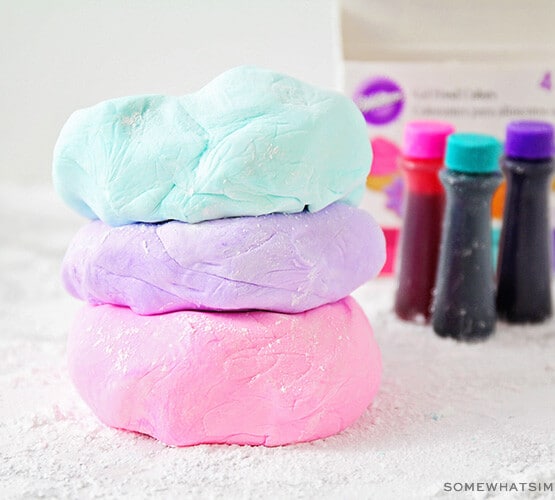three flattened balls of marshmallow fondant stacked on top of each other on a counter covered with powdered sugar. The ball of fondant on top is light blue, the middle one is light purple and the bottom one is light pink. In the background are three bottles of Wilton gel food coloring.